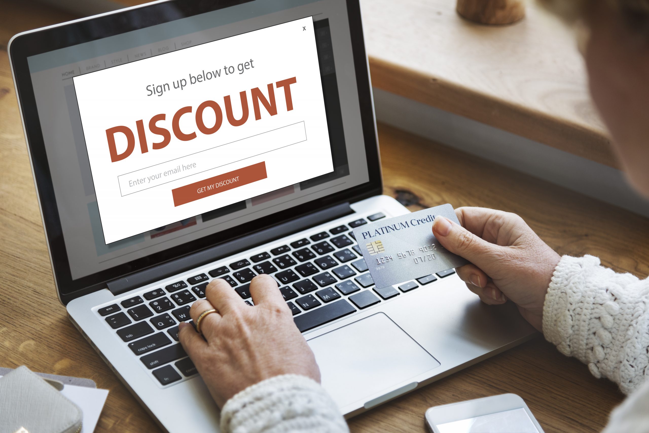 Person wearing a white sweater holding a credit card and using a laptop computer to sign up for discounts